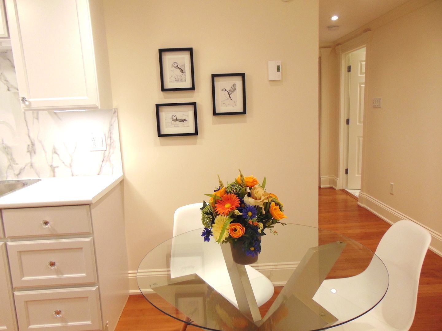 A modern room with a glass table, white chairs, a vase of flowers, and framed artwork on the wall.