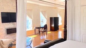 A spacious bedroom with a large bed, a television mounted on a stone wall, a desk with a chair, and a wardrobe.