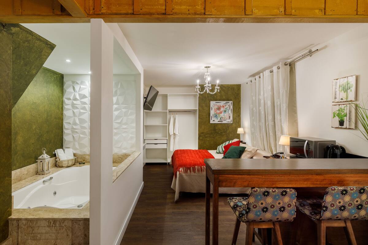 A modern hotel room with a large bathtub, a dining area with a wooden table and patterned chairs, a TV on a stand, and a chandelier.