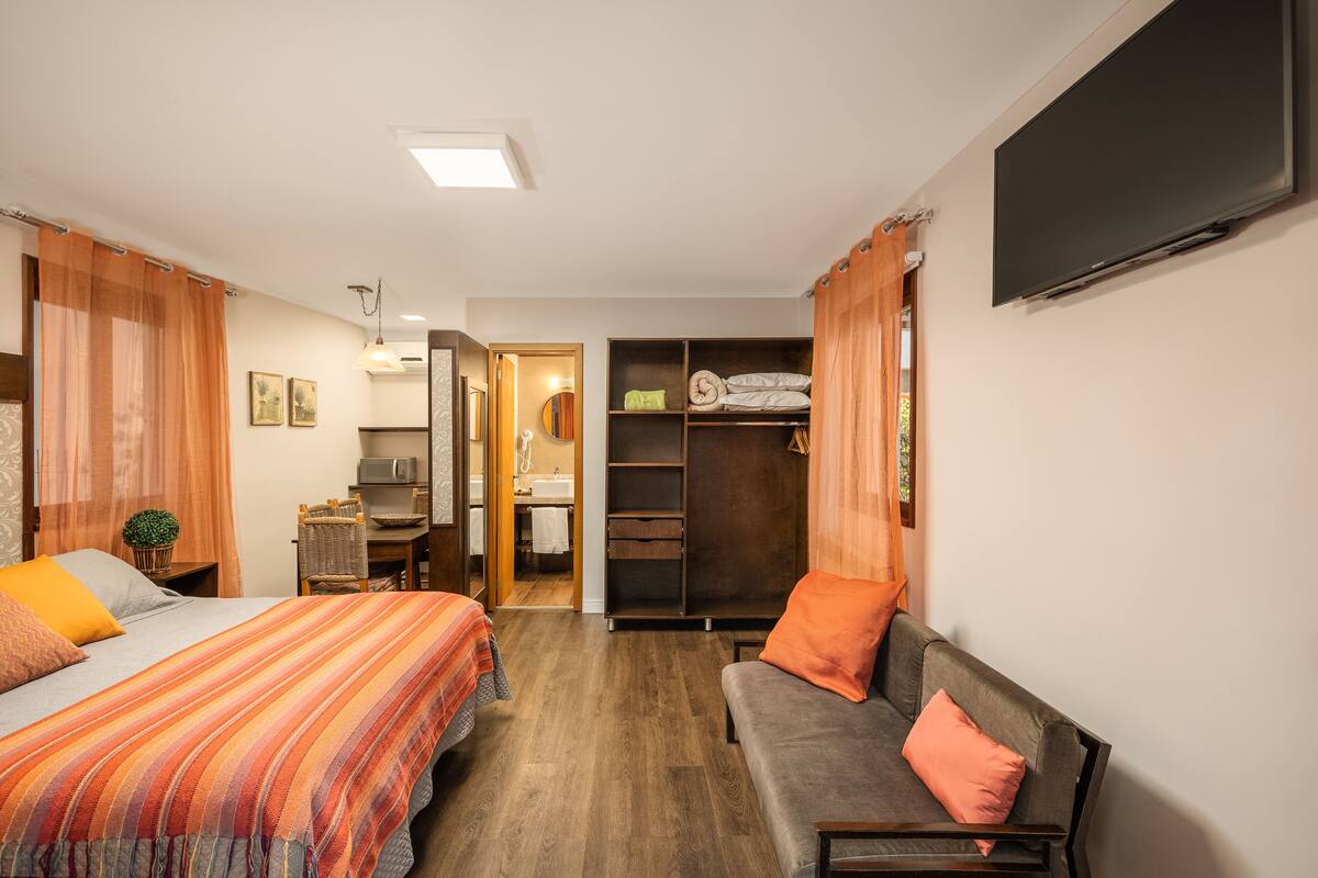 A hotel room with a bed, a sofa, a TV, a wardrobe, and a bathroom visible through an open door.