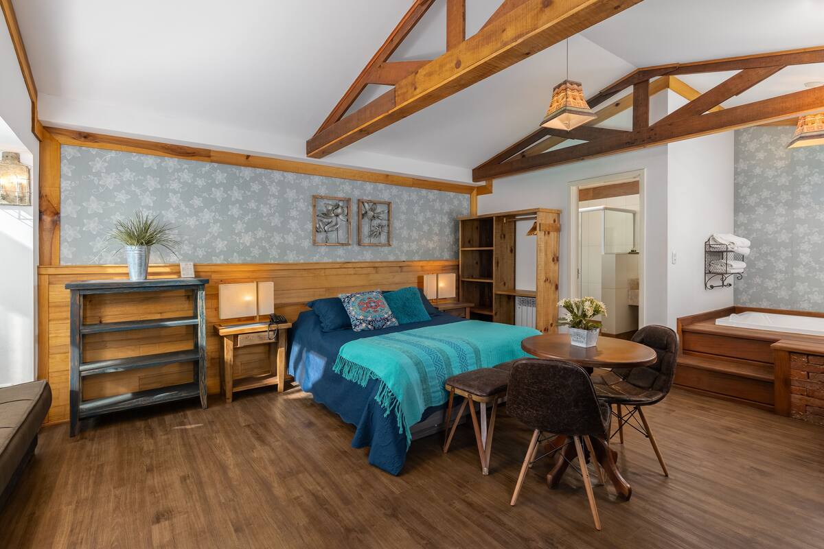 A cozy room with a bed, a small table, and a chair. There is a wooden ceiling and a window.