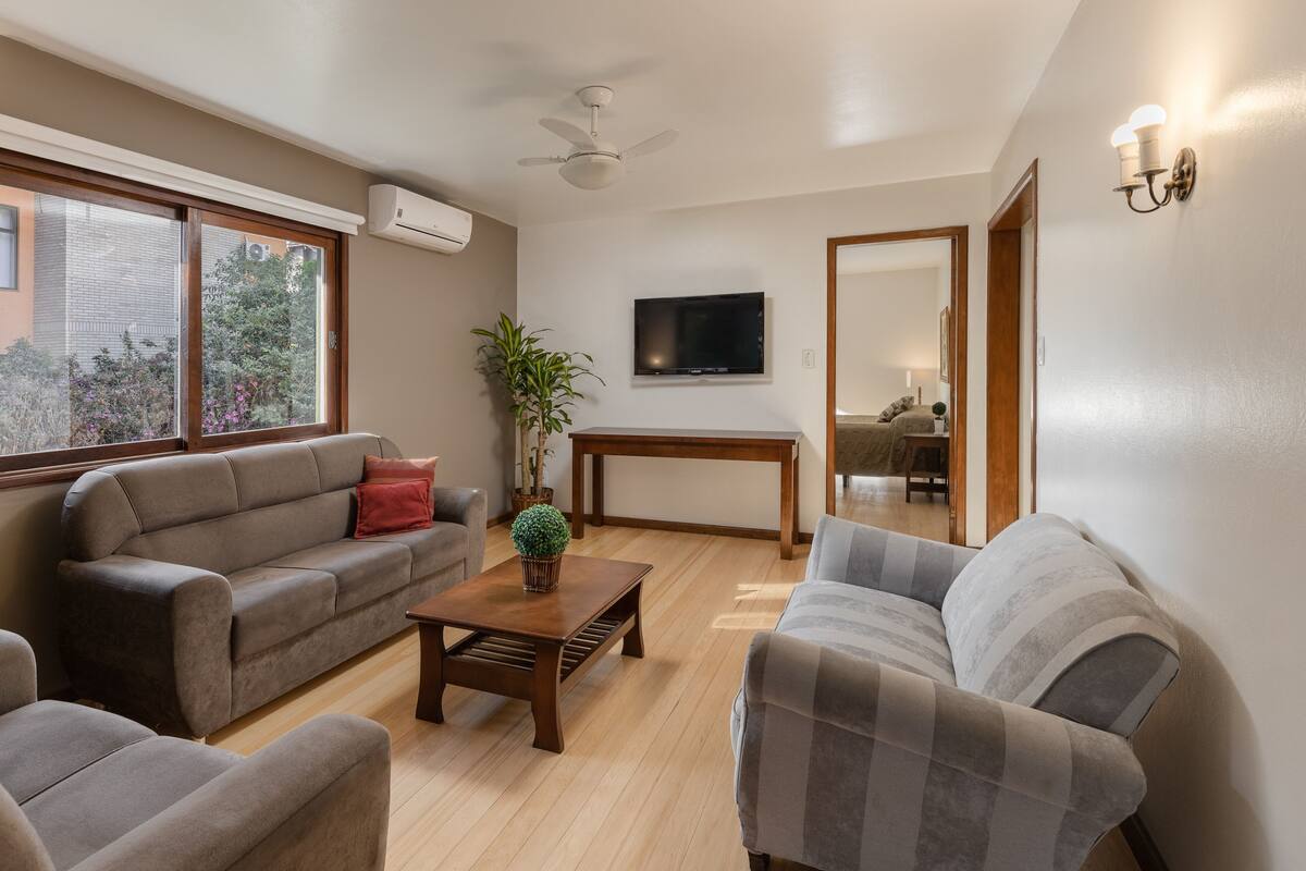 A living room with a sofa, armchair, and coffee table, a TV mounted on the wall, and a ceiling fan.