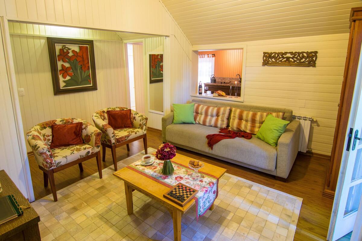 A cozy living room with floral armchairs, a sofa with colorful cushions, a wooden coffee table with a chessboard, and a vase of flowers.