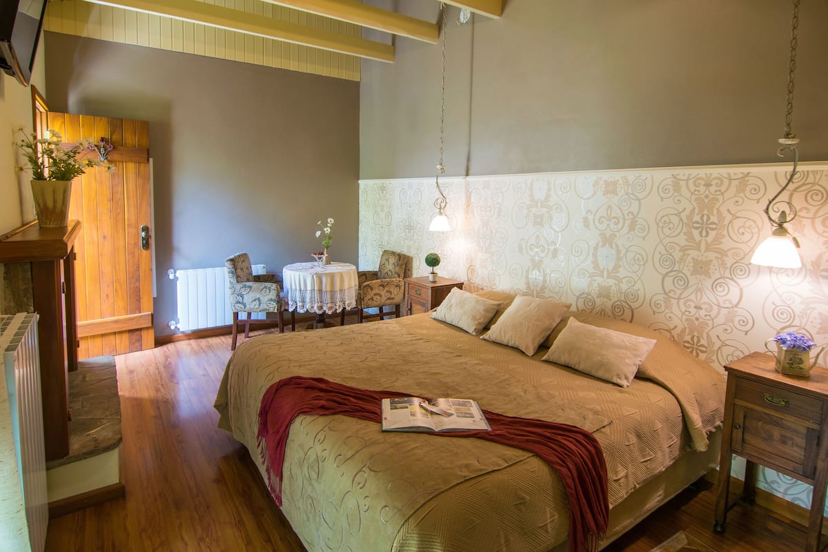 A bedroom with a large bed, a bedside table with a lamp, a chair, and a small table with a vase of flowers.