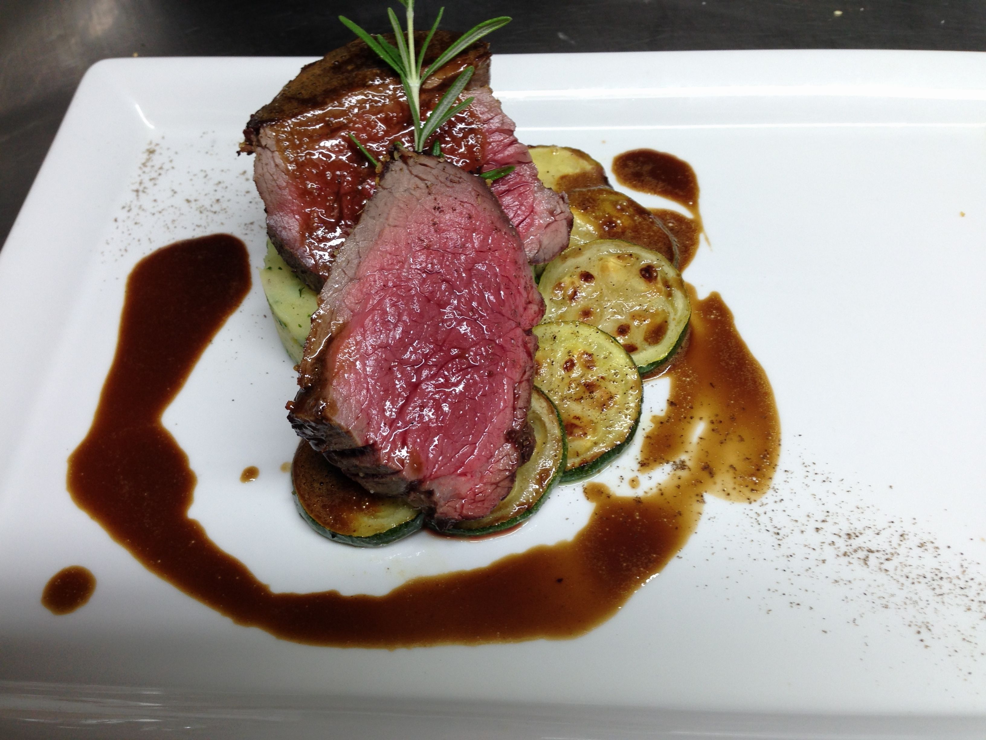 A plate of food with sliced meat, zucchini, and a sprig of rosemary.