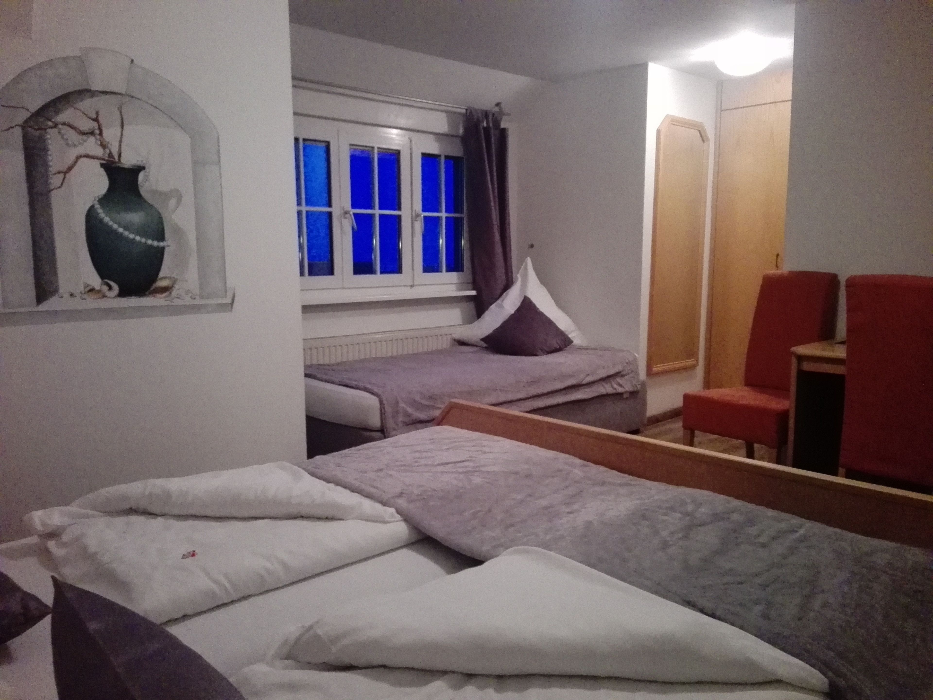 Un dormitorio con dos camas individuales, una mesita pequeña, dos sillas, una ventana con cortinas azules y un florero fijado a la pared.