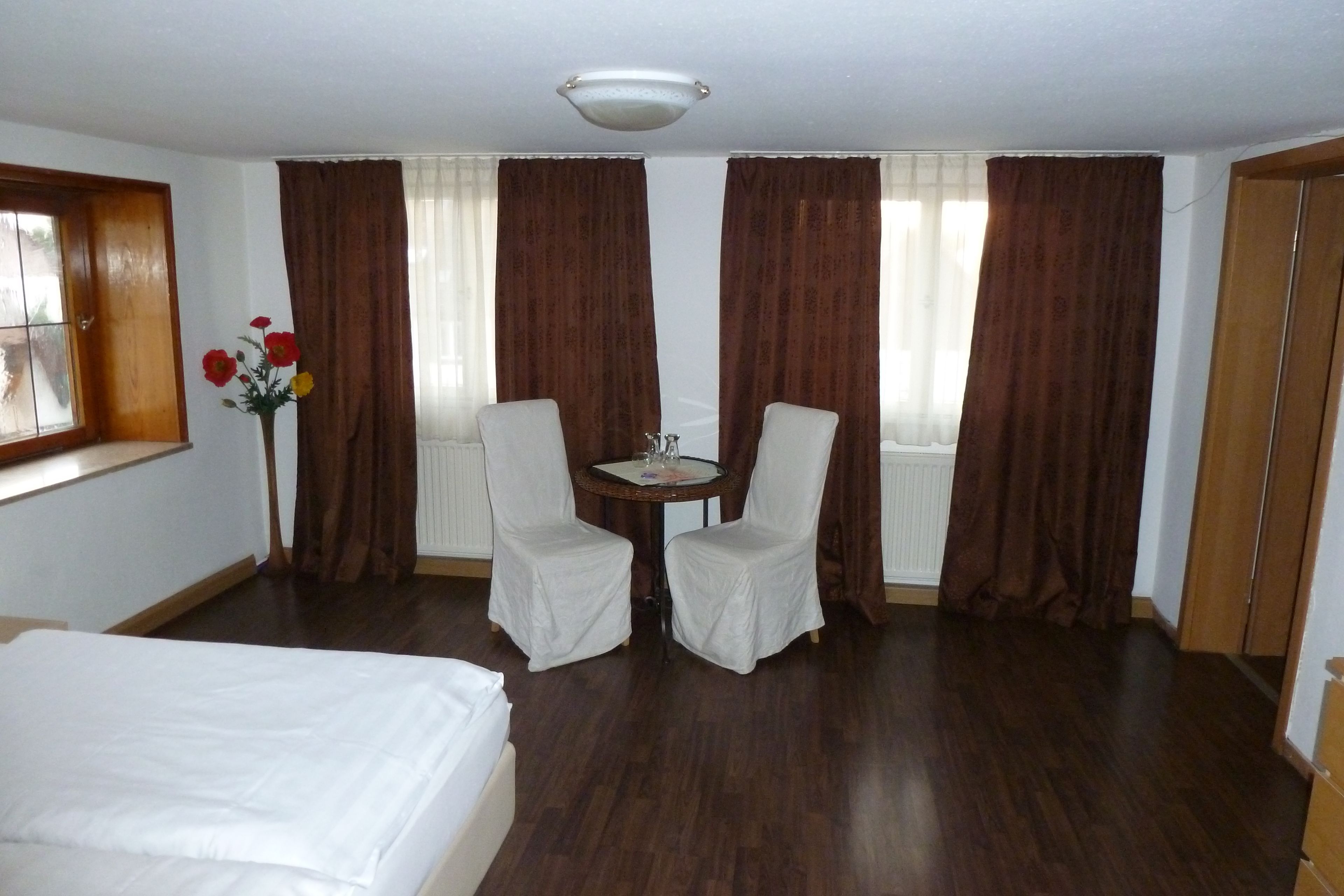 Habitación de hotel con cama, dos sillas alrededor de una mesita, un jarrón con flores y ventanas amplias con cortinas.