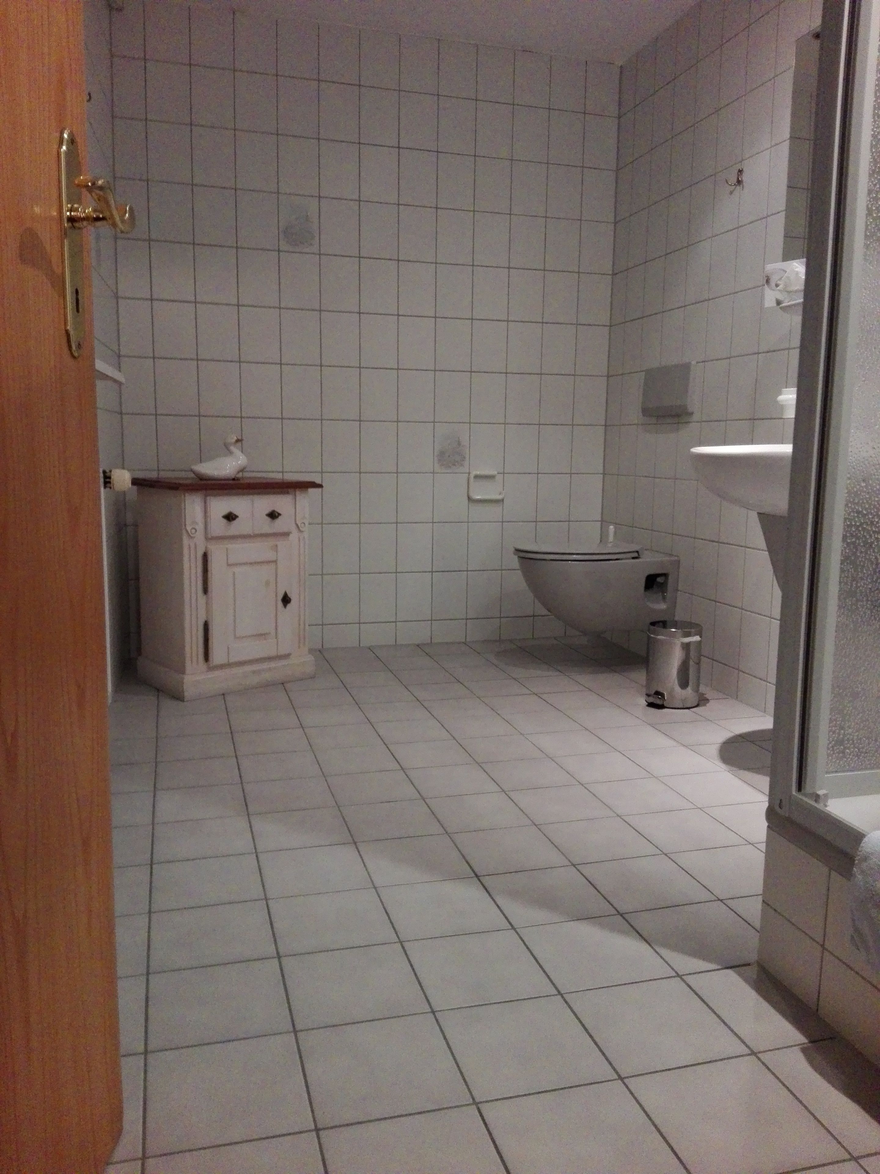 A bathroom with white tiled walls and floor, a white sink, a toilet, and a small cabinet with a decorative item on top.
