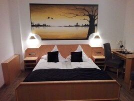 Quarto de hotel com uma cama grande, duas luminárias de cabeceira, uma escrivaninha com cadeira e um quadro com pôr do sol sobre um lago.