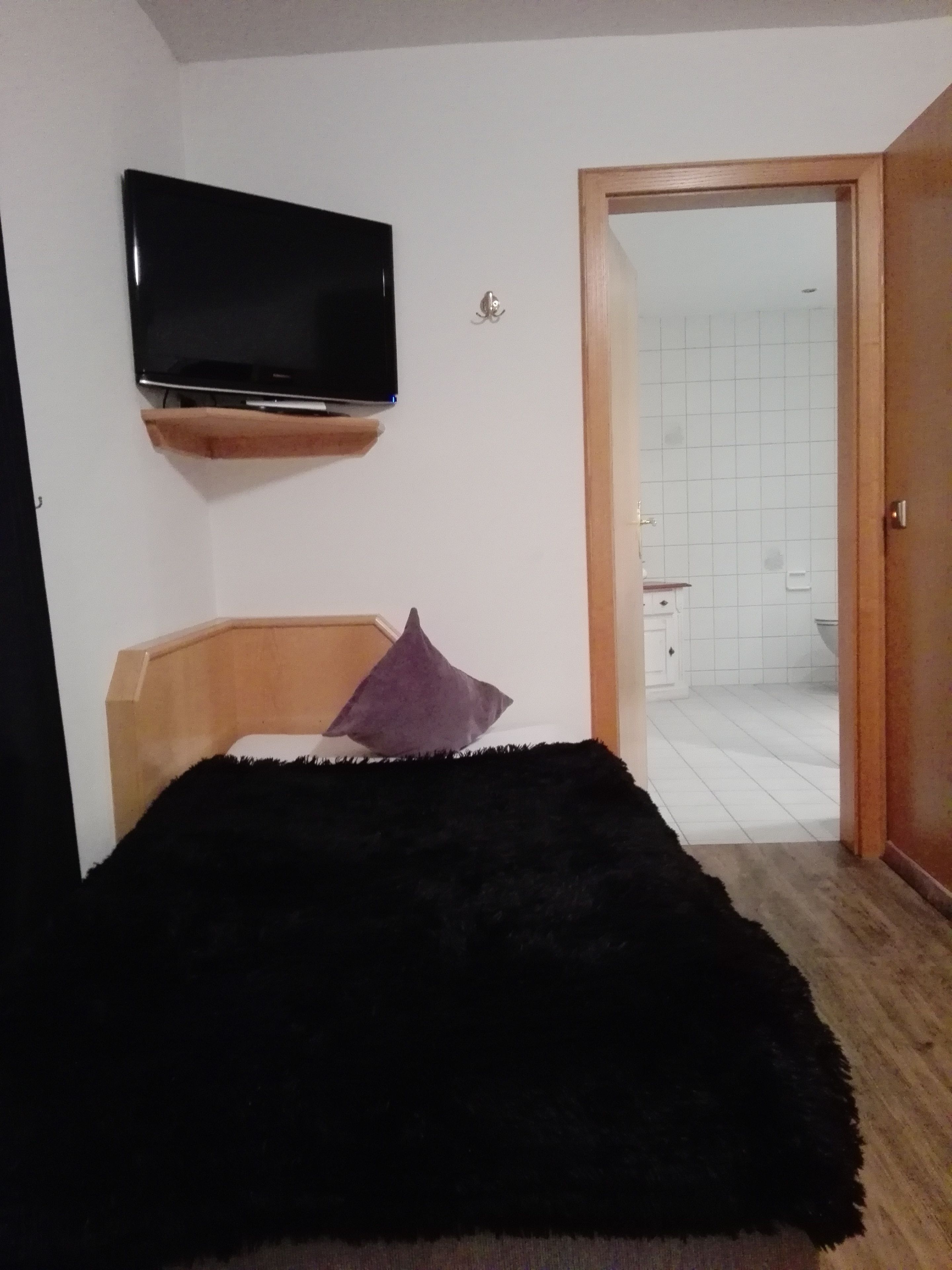 A bedroom with a bed, a television mounted on the wall, and a bathroom visible through an open door.