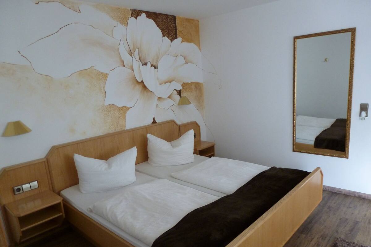 Habitación de hotel con una cama grande, cabecero de madera, ropa de cama blanca, una mesita de noche, una lámpara de noche, un espejo grande y un adorno floral en la pared.
