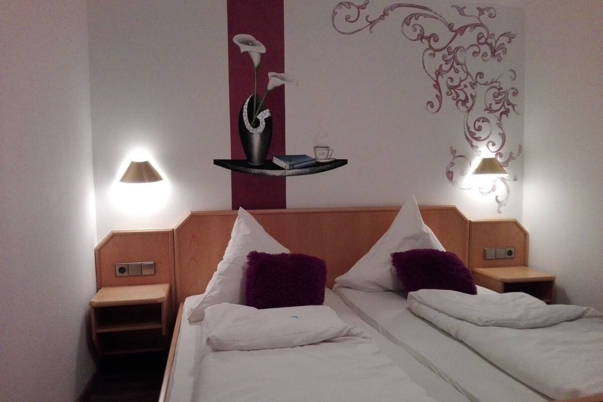 Habitación de hotel con dos camas, una mesita de noche con lámpara, un adorno en la pared y una repisa con taza y platillo.