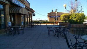 Une terrasse avec des sièges extérieurs, et en arrière-plan, un bâtiment portant les inscriptions « CAFÉTÉRIA » et « HÔTEL SOL ».