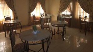 A dining area with round tables and chairs, a kitchen area, and large windows with curtains. - New Gate Lodge & Hospitality Ltd (Abuja)