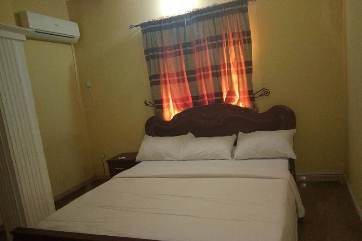 Una habitación de hotel estándar con una cama individual, una mesita de noche, un equipo de aire acondicionado y una ventana con cortinas a cuadros.