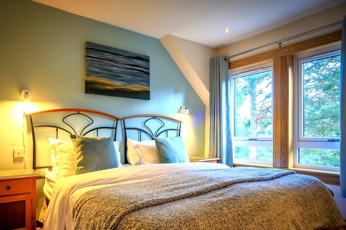 Une chambre à coucher avec un lit, des tables de chevet, un tableau et de grandes fenêtres donnant sur la forêt.
