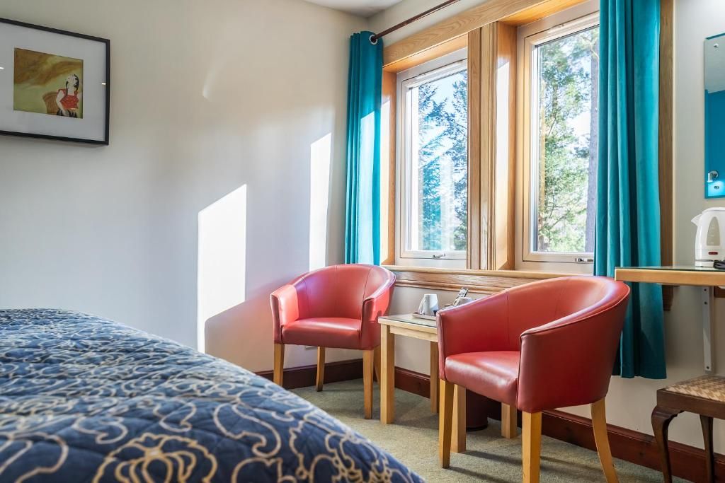 Une chambre d’hôtel avec un lit, deux chaises rouges, une petite table, une fenêtre avec des rideaux et un tableau au mur.