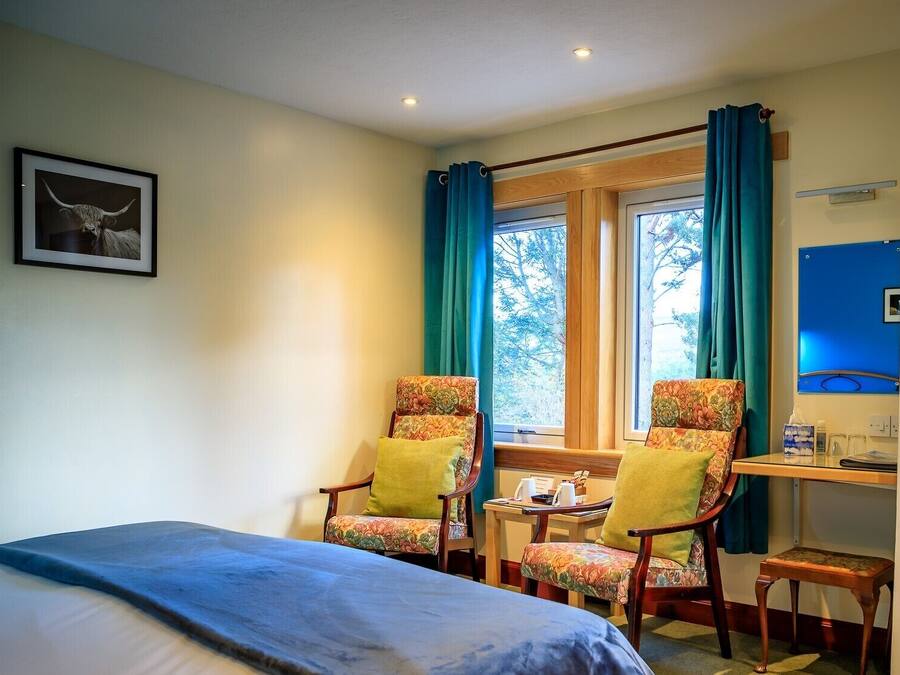 A room with a bed, two floral armchairs, a small table with a lamp, and a window with curtains.
