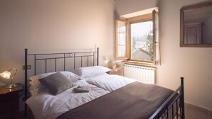 A neatly made bed with white linens and a brown blanket, a bedside table with a lamp, and a window with a view of trees and a building. - Buccia Nera (Arezzo)