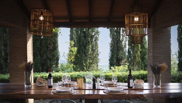An outdoor dining area with a wooden table set for two, surrounded by chairs, and two hanging light fixtures. - Buccia Nera (Arezzo)