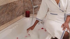 A person in a white bathrobe sitting in a bathtub with rose petals, a red candle, and a marble wall in the background. - Auberge le Rajah (Quatre Bornes)