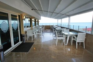A covered outdoor dining area with white chairs and tables, a black mat at the entrance, and a view of the sea. - Green Suada Hotel (Pazar)