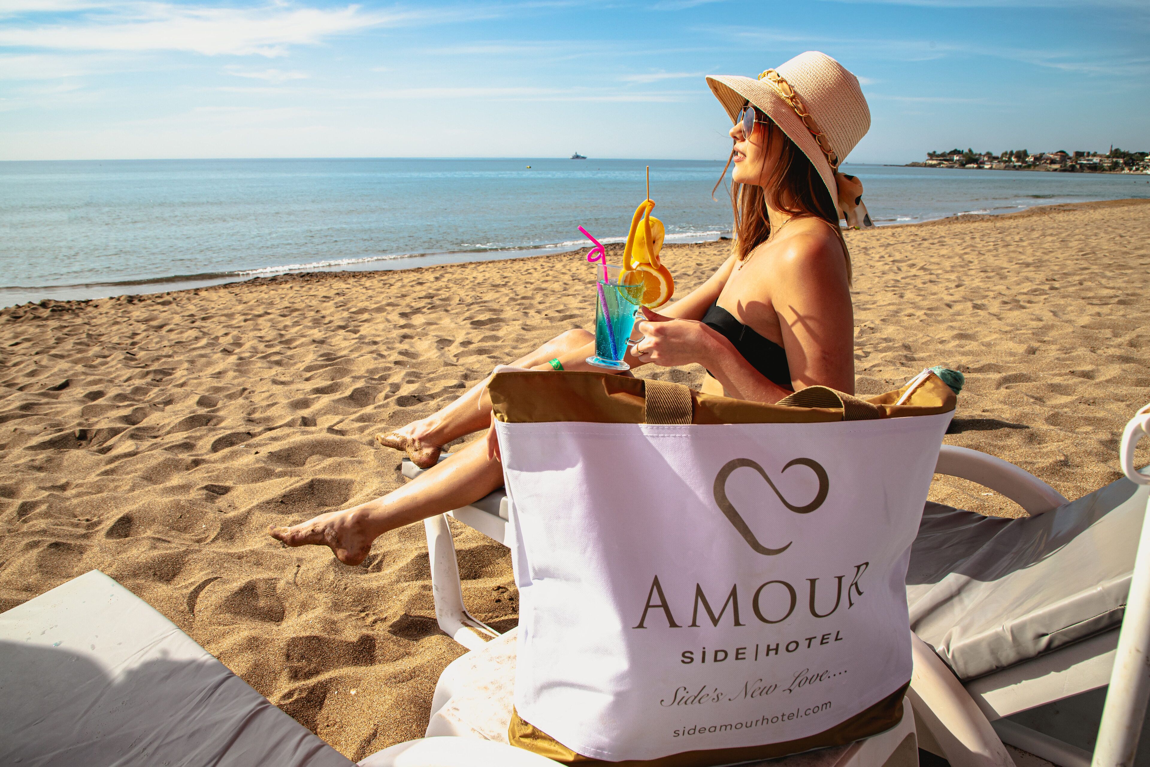 Una persona sentada en una silla de playa con un bolso de playa con la etiqueta "AMOUR SIDEHOTEL" y una bebida con pajita y una rodaja de naranja.