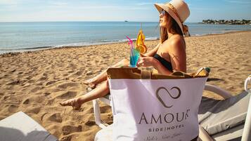 Una persona sentada en una silla de playa con un bolso de playa con la etiqueta "AMOUR SIDEHOTEL" y una bebida con pajita y una rodaja de naranja.