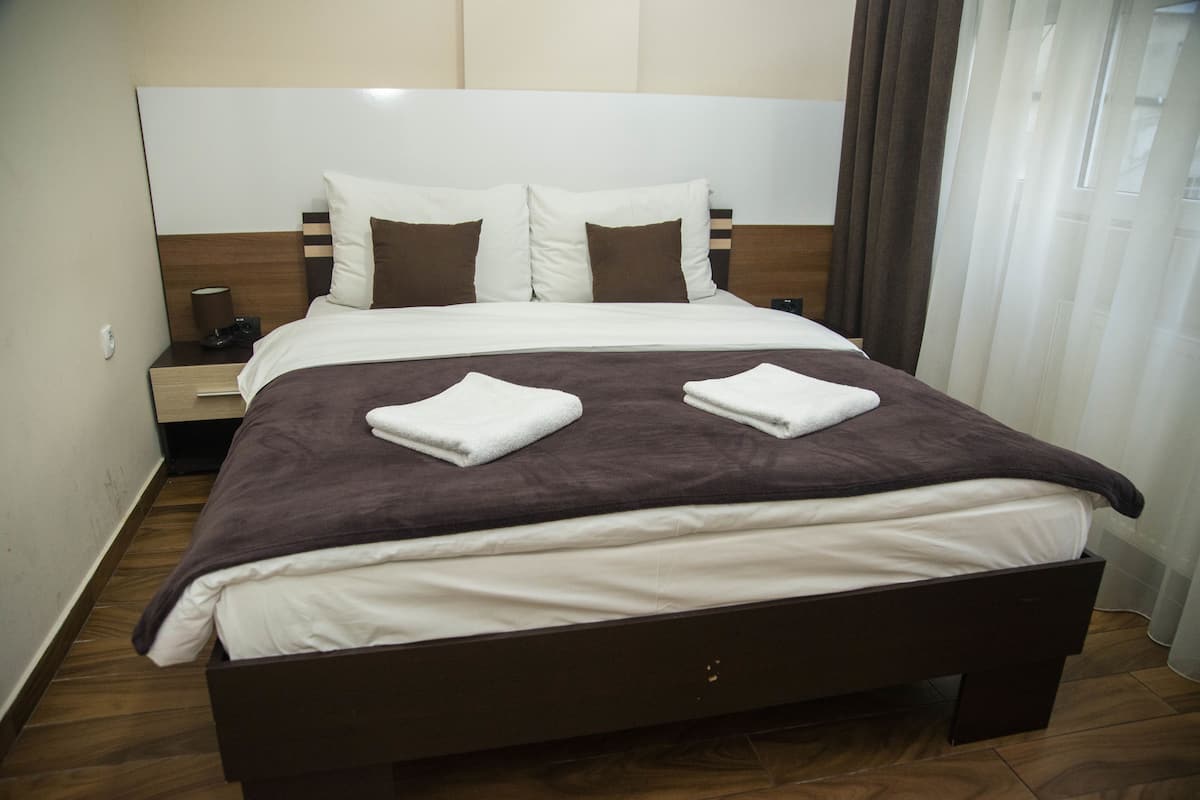 Cama bem arrumada com cabeceira marrom, roupa de cama branca e duas toalhas brancas dobradas.