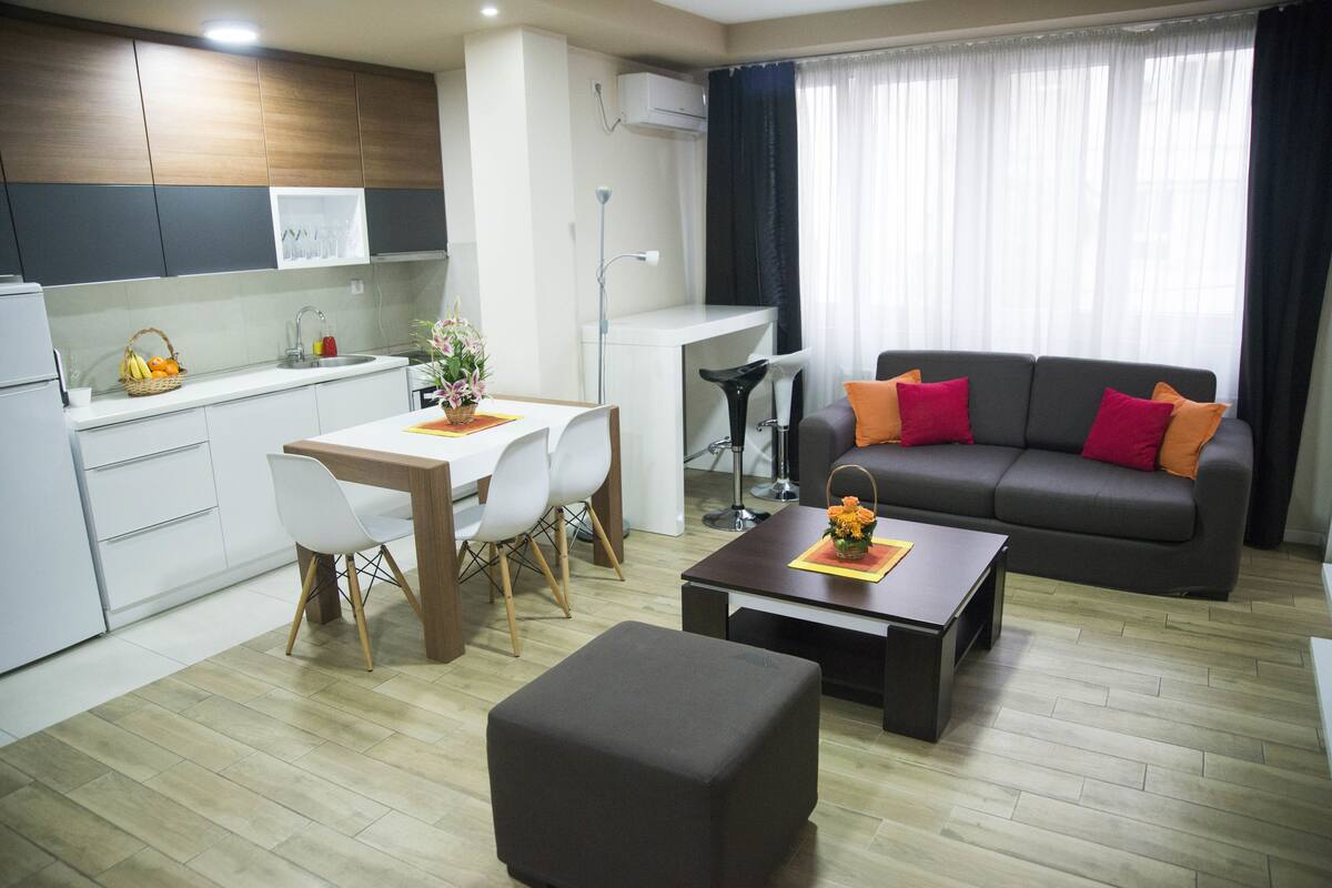 Uma sala de estar moderna com um sofá cinzento, uma mesa de centro e uma área de jantar com uma mesa e cadeiras brancas.
