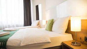 Habitación de hotel con dos camas, una manta verde, almohadas blancas, una lámpara de noche y una ventana con cortinas.