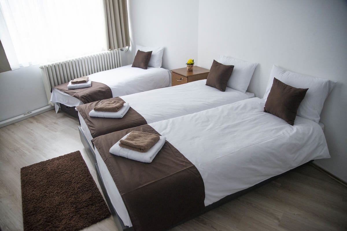Quarto de hotel com duas camas de solteiro, uma mesa de cabeceira de madeira, um tapete castanho e uma janela com cortinas.