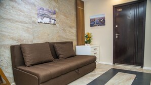 Un sofá marrón con dos cojines, una mesita auxiliar blanca con un florero, una puerta de madera oscura con el número 100, y dos cuadros enmarcados en la pared.
