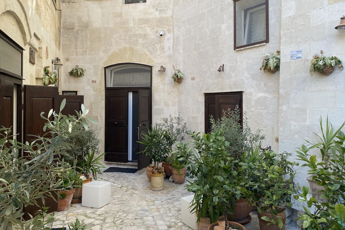 Une cour avec un sol en pierre, une porte centrale et des plantes en pot.