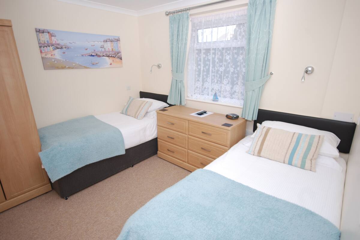 A bedroom with two beds, a dresser, a window with curtains, and a painting on the wall.