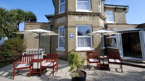 A two-story building with a corner turret, a patio with wooden chairs and umbrellas, and a small potted plant. - Westbury Lodge (Shanklin)