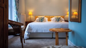 A hotel room with a bed, two bedside lamps, a wicker chair, a small round table, and a large mirror on the wall. - Westbury Lodge (Shanklin)