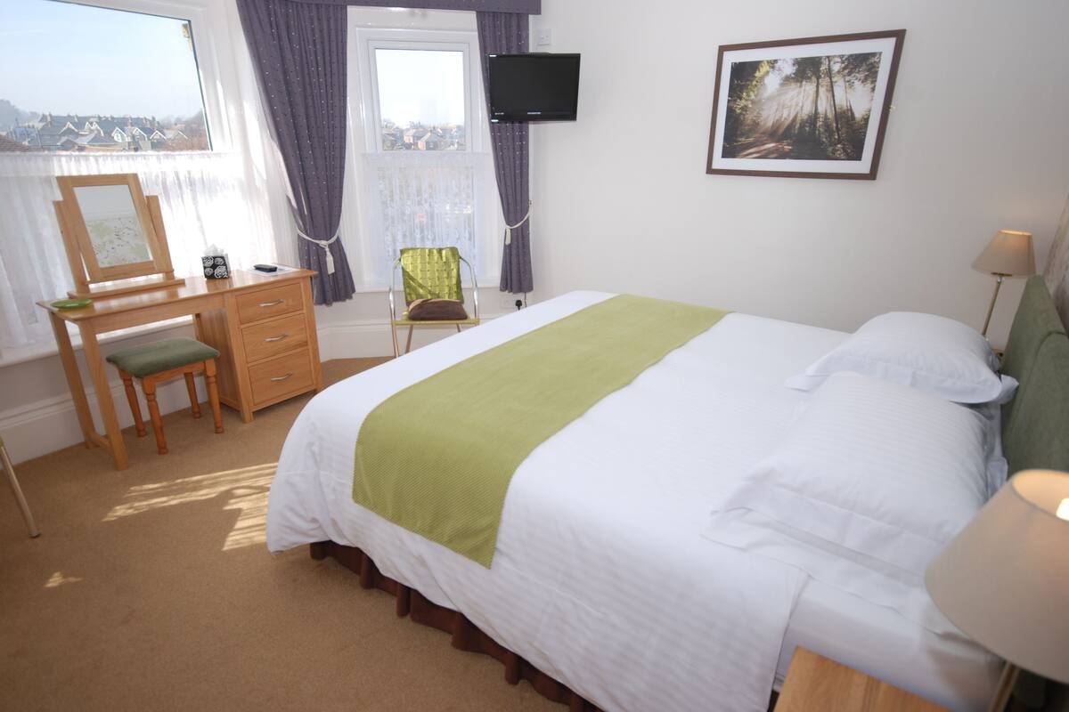 A bedroom with a bed, a desk, a chair, a television, and a window with curtains.