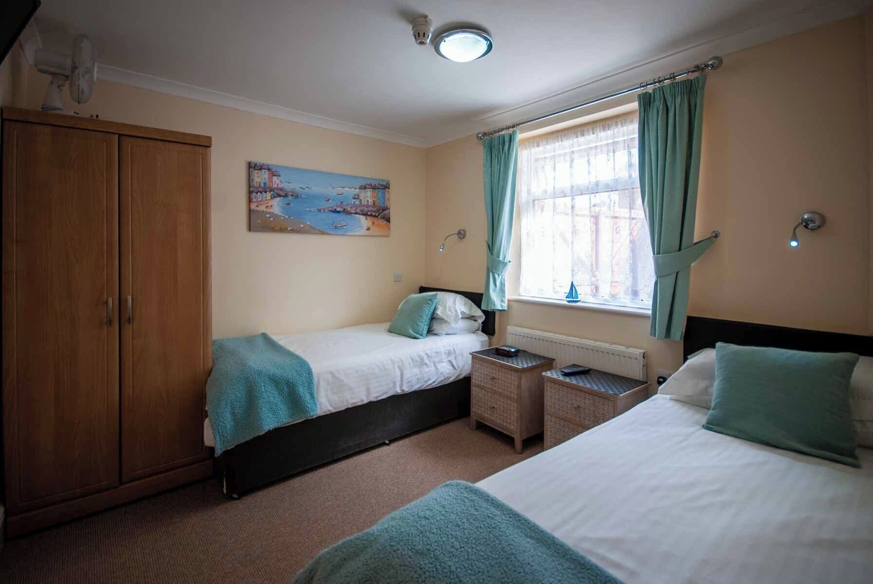 A bedroom with two beds, a wooden wardrobe, a painting of a coastal scene, a window with curtains, and a nightlight.