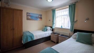 A bedroom with two beds, a wooden wardrobe, a painting of a coastal scene, a window with curtains, and a nightlight.