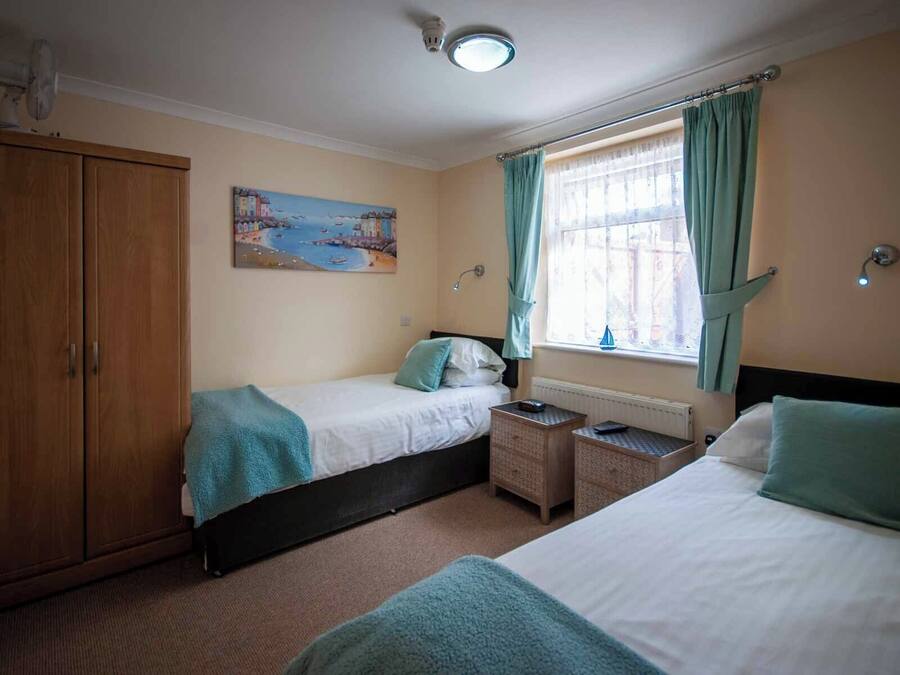A bedroom with two beds, a wooden wardrobe, a painting of a coastal scene, a window with curtains, and a nightlight.