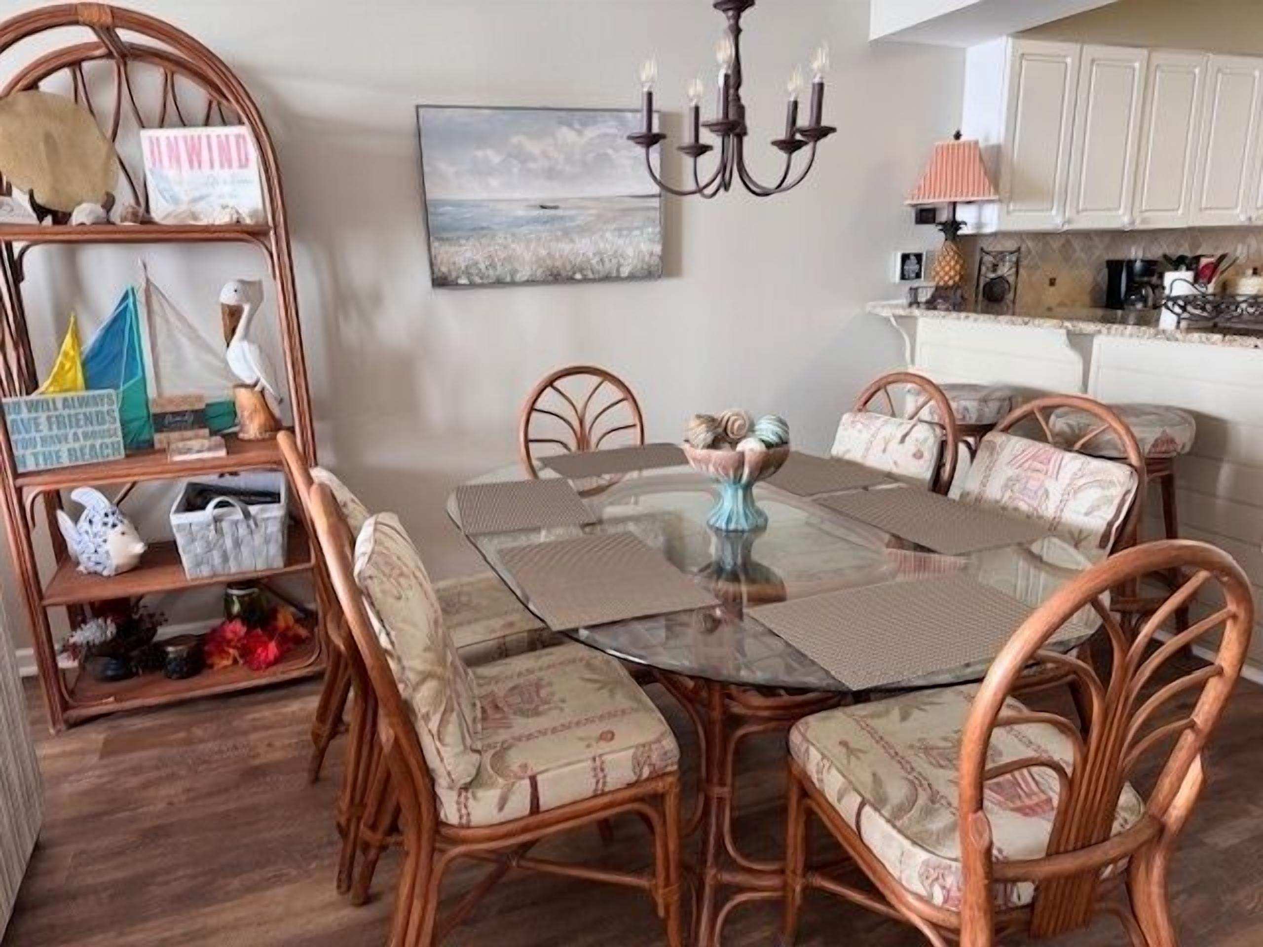 A dining area with a glass table surrounded by wicker chairs, a chandelier, and a kitchen in the background.