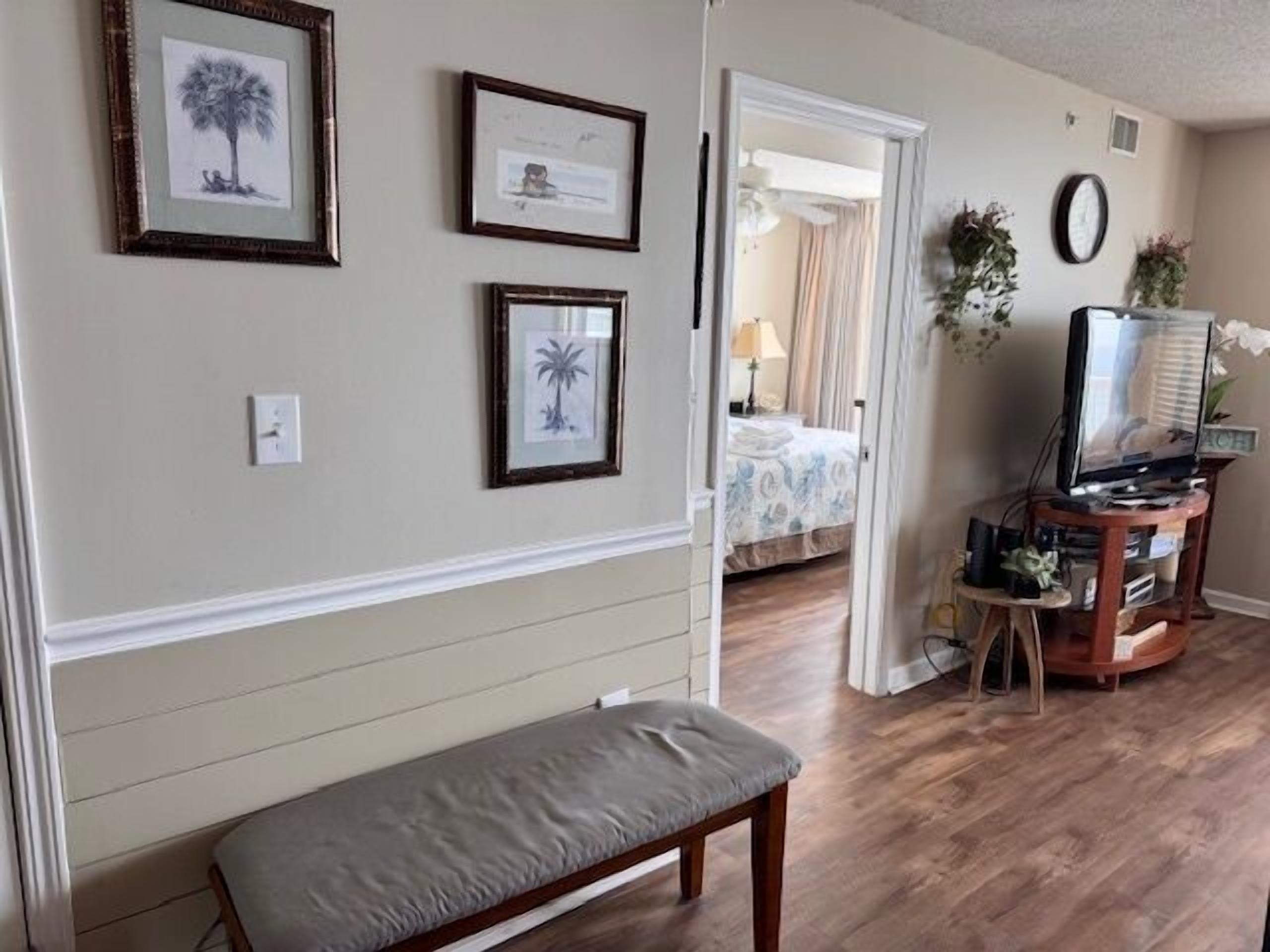 A room with a wooden bench, framed artwork on the wall, a television on a stand, and a doorway leading to another room.