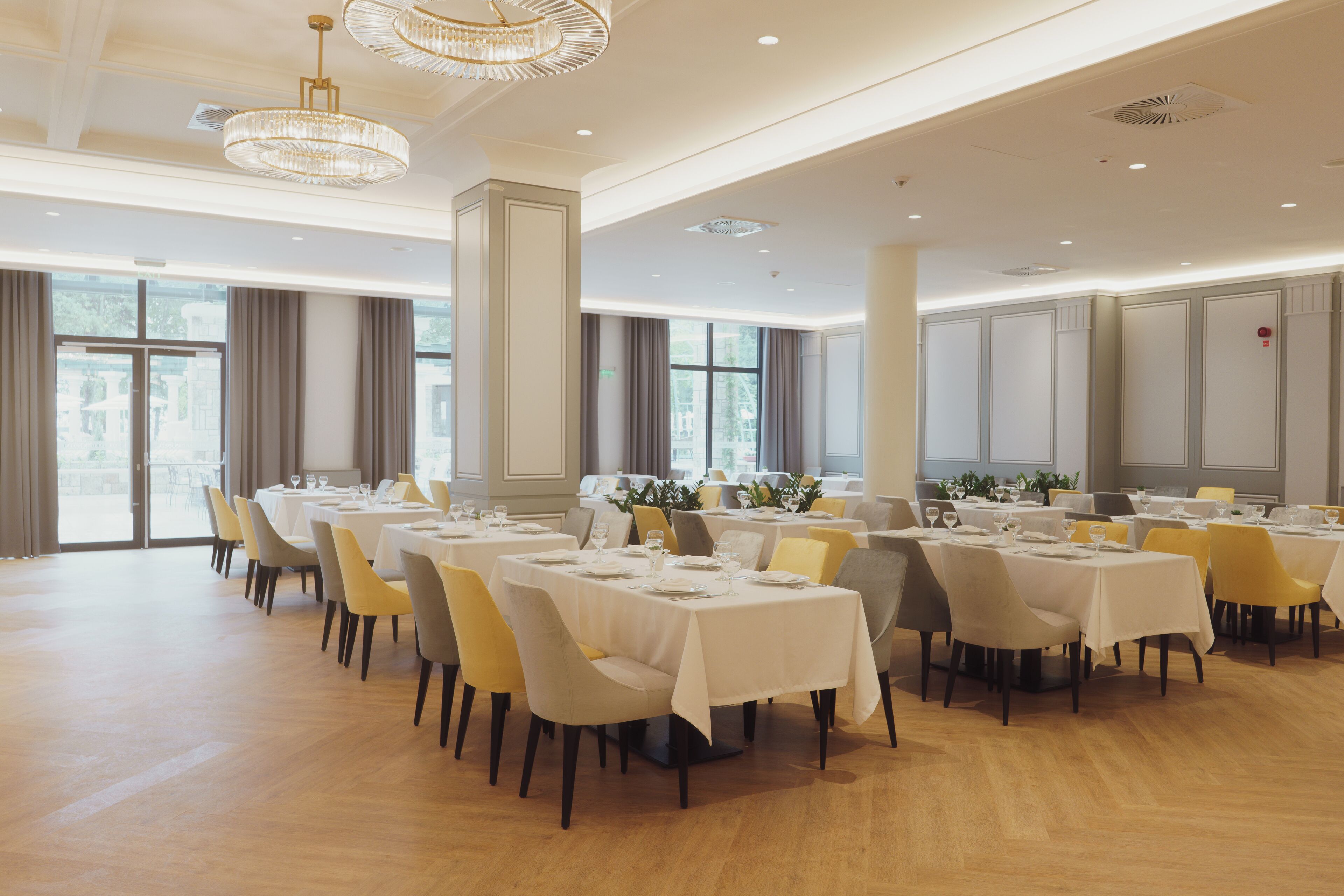 A spacious dining area with multiple tables and chairs, large windows, and elegant chandeliers.