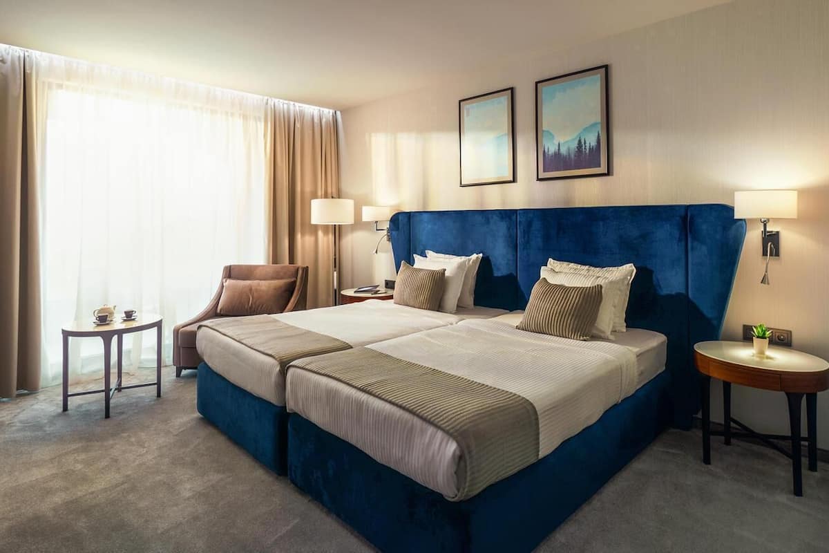 A double bed with a blue headboard, two bedside tables, a small side table, and a window with curtains.