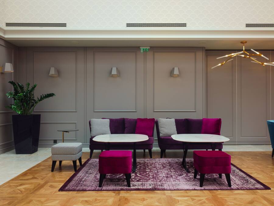 Un'area lounge moderna con due tavoli rotondi, poltrone imbottite viola e nere, una grande pianta in vaso e un tappeto decorato.