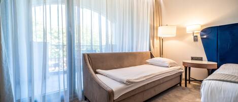 A hotel room with a bed, a small table, a lamp, and a view through sheer curtains.