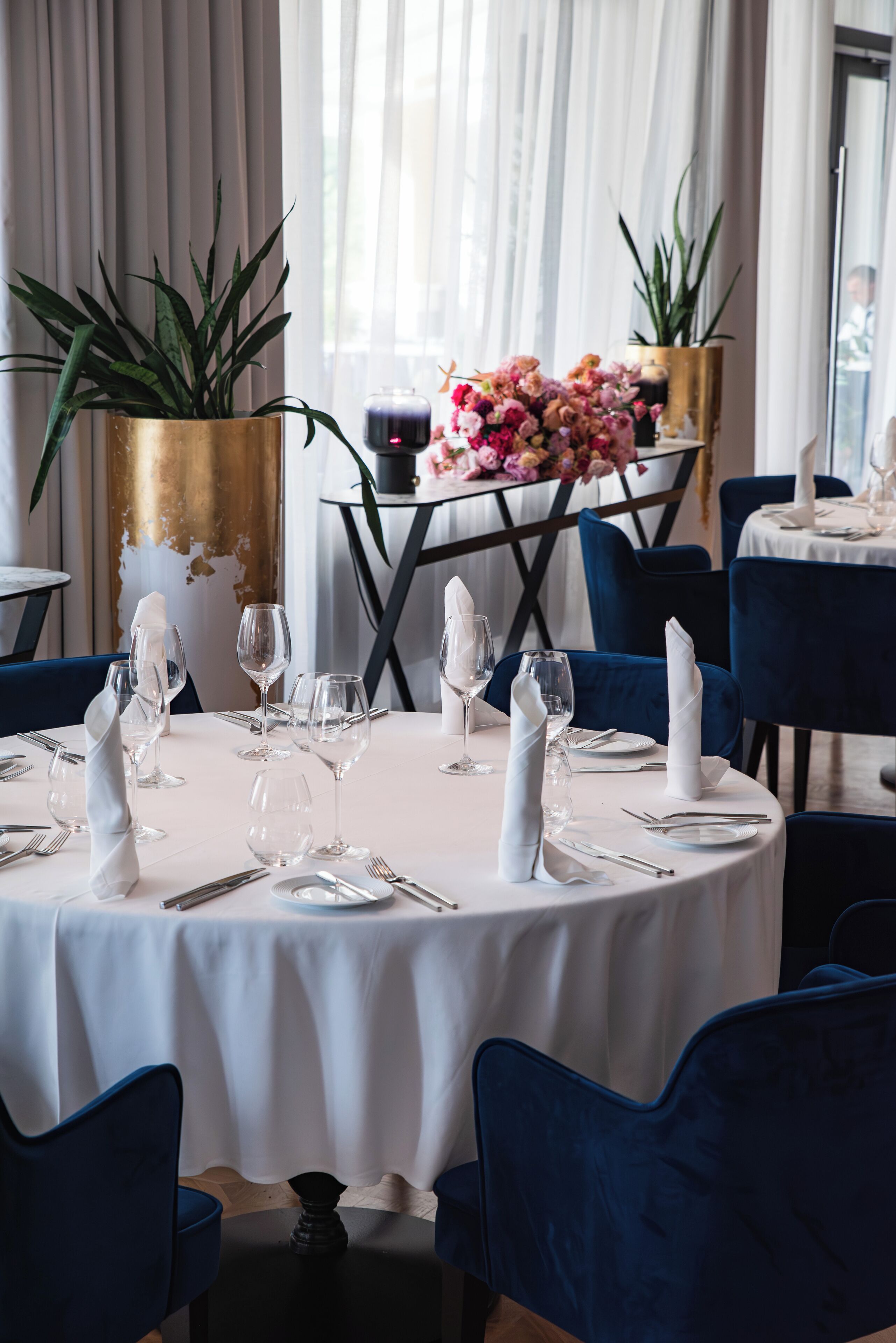 A round dining table set with white tablecloth, blue chairs, and a vase of pink flowers.