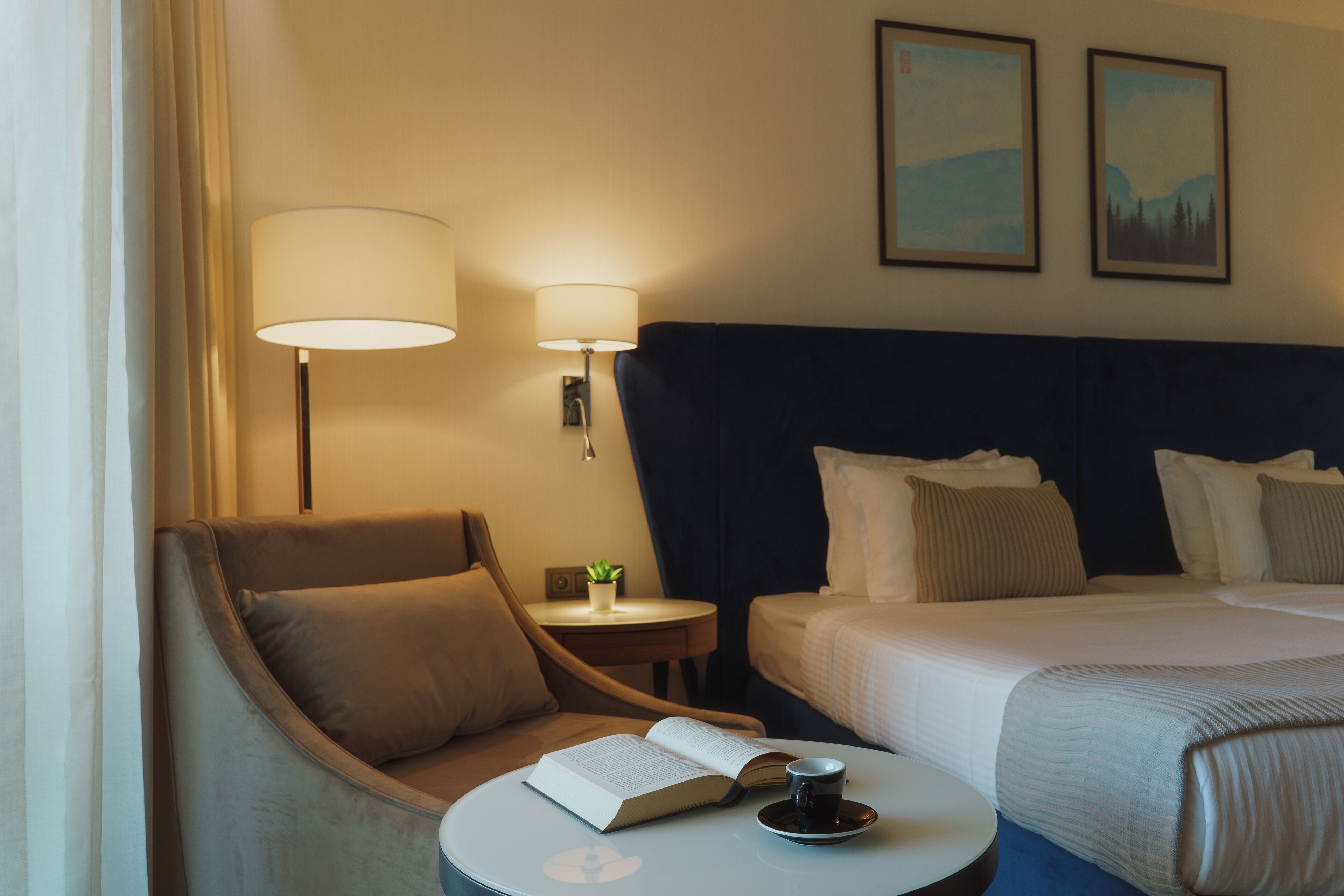 A hotel room with a bed, a chair, a small table with a book and a cup, two lamps, and two framed pictures on the wall.
