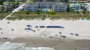 An aerial view of a beach with a large building complex in the background, including a pool area.